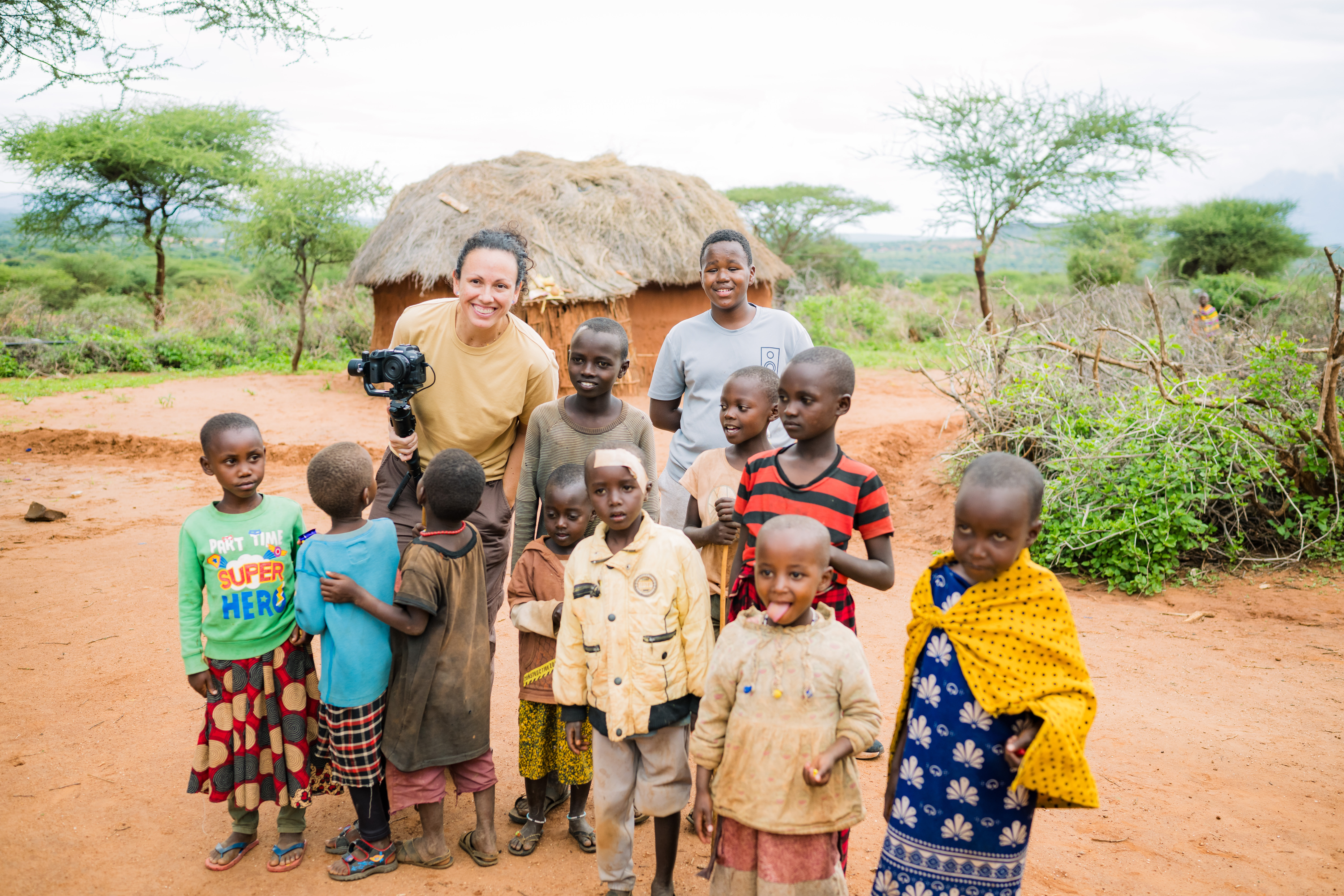 Woman with camera posing with a group of Maasai children in a rural village setting, highlighting community engagement and support for Family Village Foundation's mission.